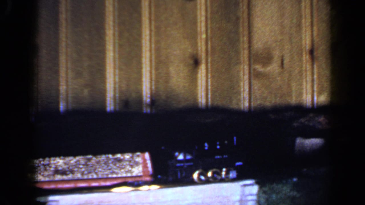 1965 Long Island home-movie frame with toy train cars moving right to left across a miniature bridge.