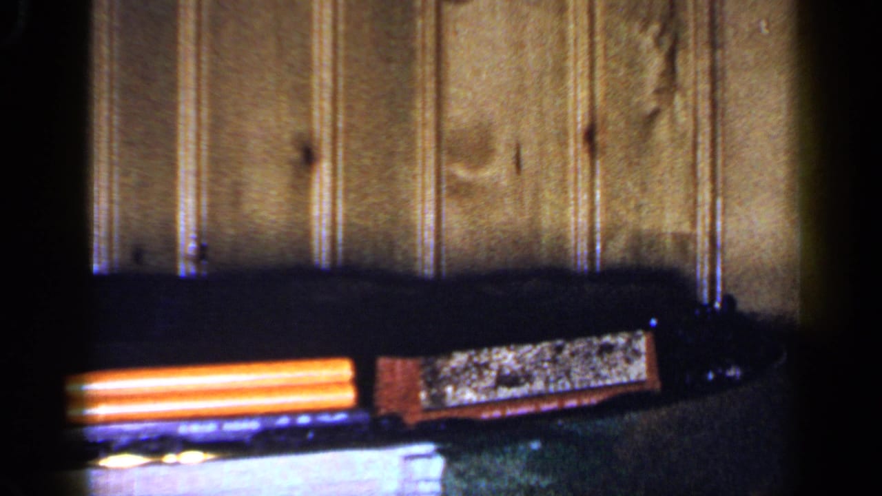 1965 Long Island home-movie frame with an orange toy freight car moving over a small bridge before a wood-paneled wall.