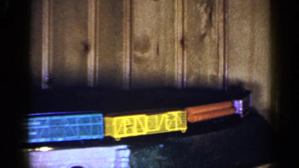 A colorful model train with blue, yellow, and orange cars travels across a dark track in this 1965 Long Island home-movie frame.