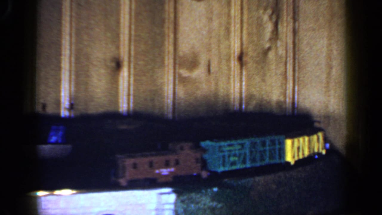The model train continues around the track, with a darker engine area and green-yellow cars visible against wood paneling.