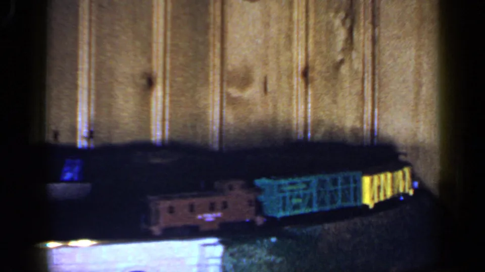 The model train continues around the track, with a darker engine area and green-yellow cars visible against wood paneling.