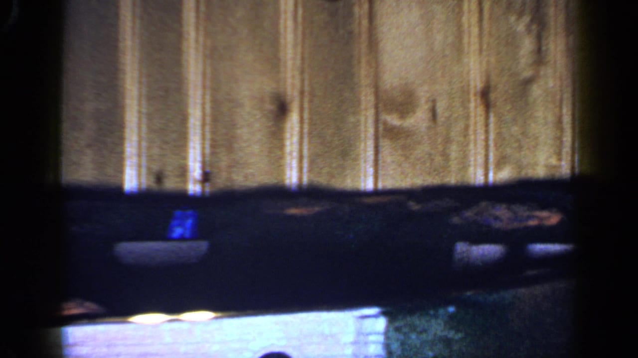 1965 Long Island home-movie frame showing a dark toy train on curved indoor track against wood-paneled walling.