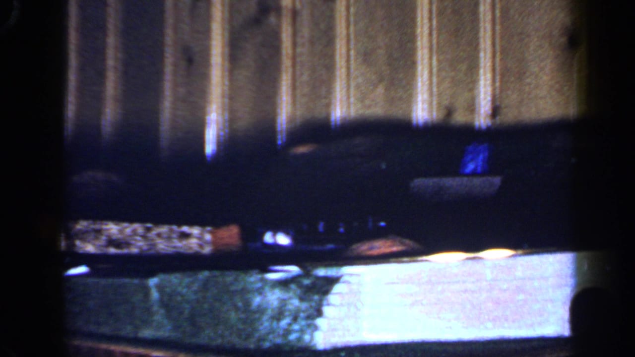 The toy train and cars sit or pass along the track, with a small blue object and wood paneling visible in the 8mm frame.