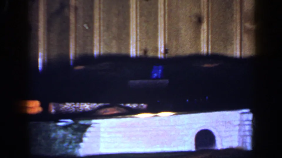 1965 Long Island 8mm frame showing a dim interior edge, bright lower surface, and small arched object near a wood-paneled wall.