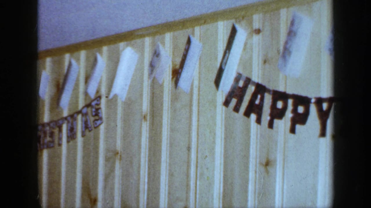 1965 Long Island home-movie frame showing paper decorations and a HAPPY BIRTHDAY banner on a wood-paneled interior wall.