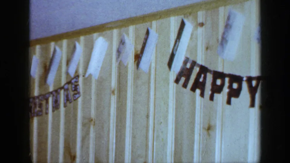1965 Long Island home-movie frame showing paper decorations and a HAPPY BIRTHDAY banner on a wood-paneled interior wall.