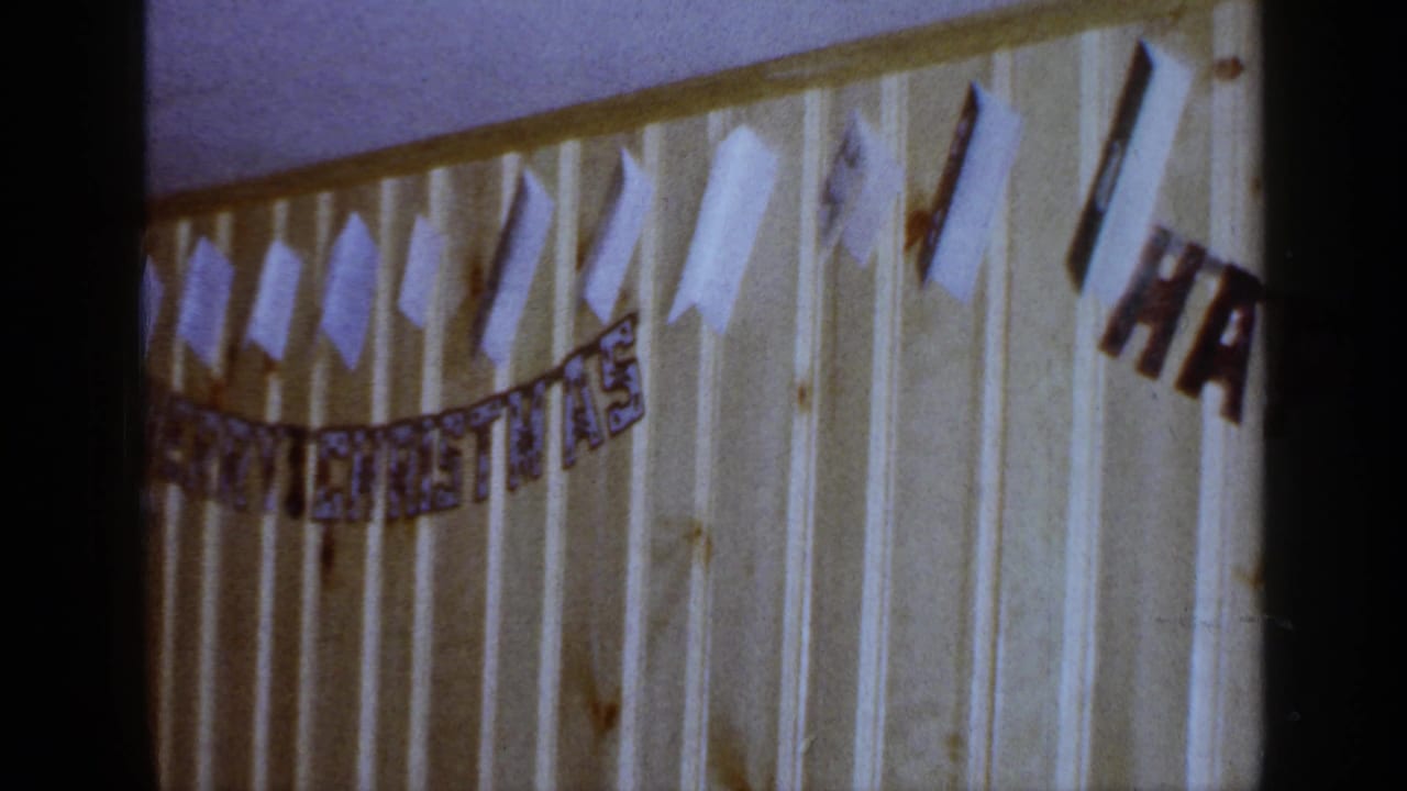 1965 Long Island home-movie frame showing a MERRY CHRISTMAS banner and taped white cutouts on a pale paneled wall.