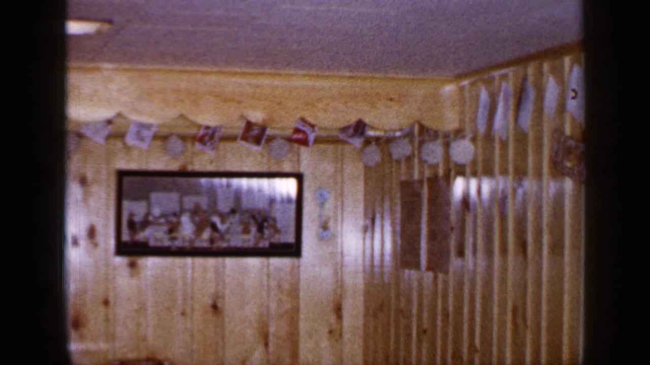 1960s home-movie frame showing a framed gathering photograph below Christmas garland in a wood-paneled Long Island interior.