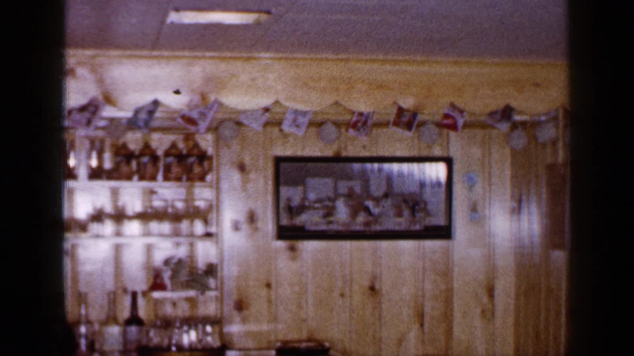 1965 Long Island home-movie frame showing a wood-paneled bar area with bottles, glassware, a framed picture, and small flags.