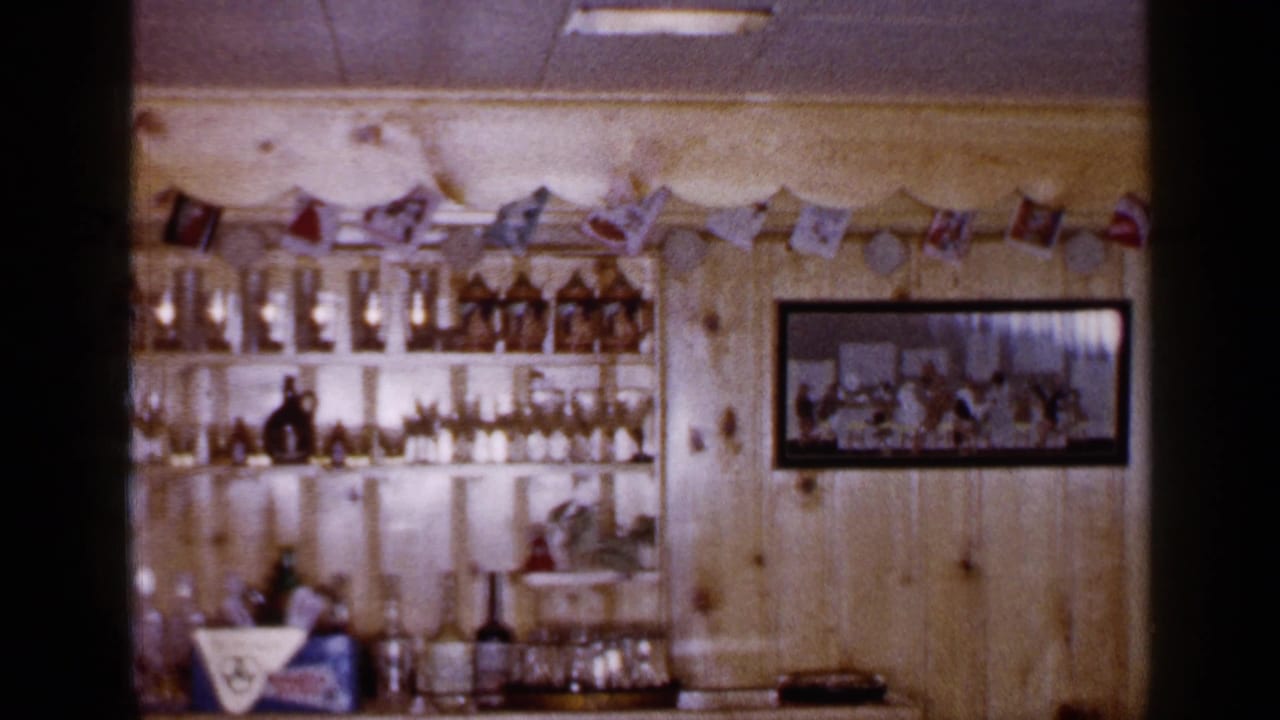 1965 Long Island interior with stocked bar shelves, glassware, wood paneling, flag garland, and a framed picture on the wall.