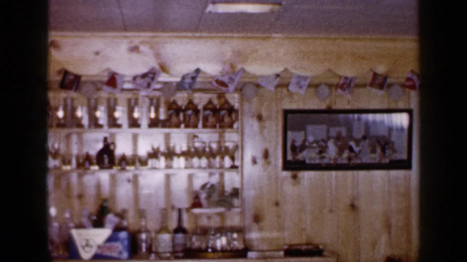 1965 Long Island interior with stocked bar shelves, glassware, wood paneling, flag garland, and a framed picture on the wall.