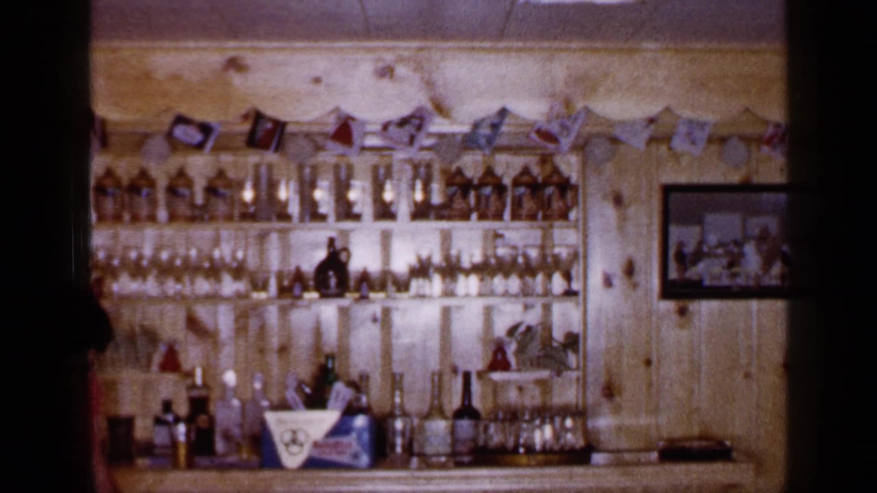 1965 Long Island home bar view with wood paneling, rows of bottles, glassware, a counter, flag garland, and wall decor.