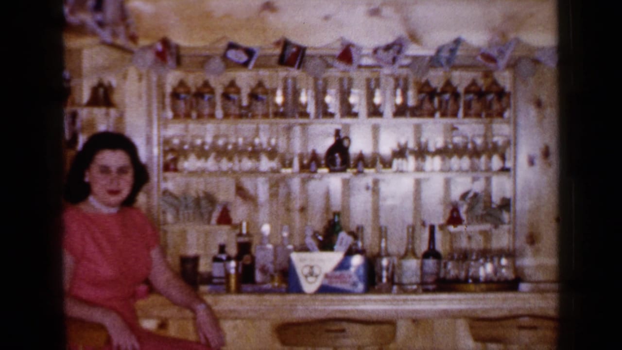 1965 Long Island home-movie frame of a woman in a red dress seated at a wood-paneled bar stocked with bottles and glassware.