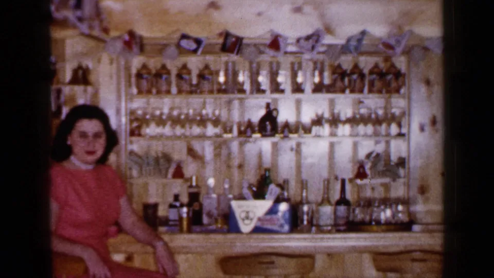 1965 Long Island home-movie frame of a woman in a red dress seated at a wood-paneled bar stocked with bottles and glassware.
