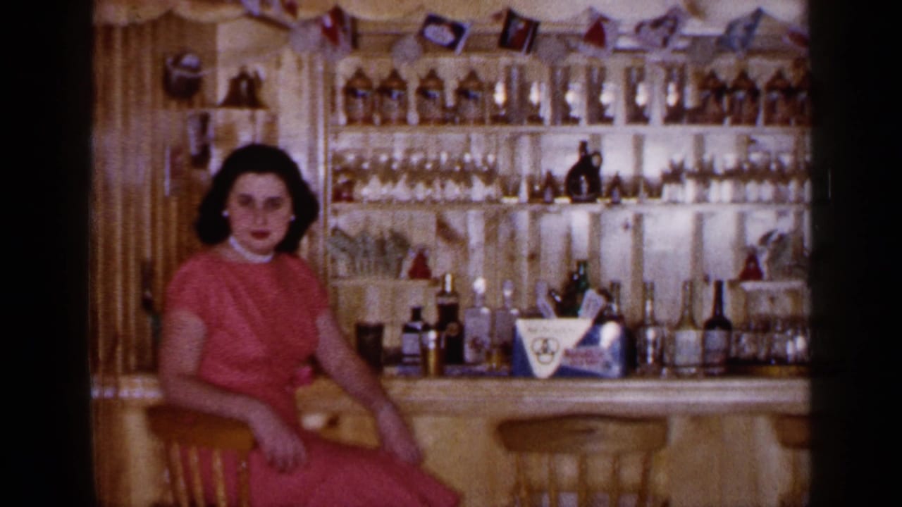 Dark-haired woman in a pink dress sitting at a wooden home bar with bottles and glassware behind her.