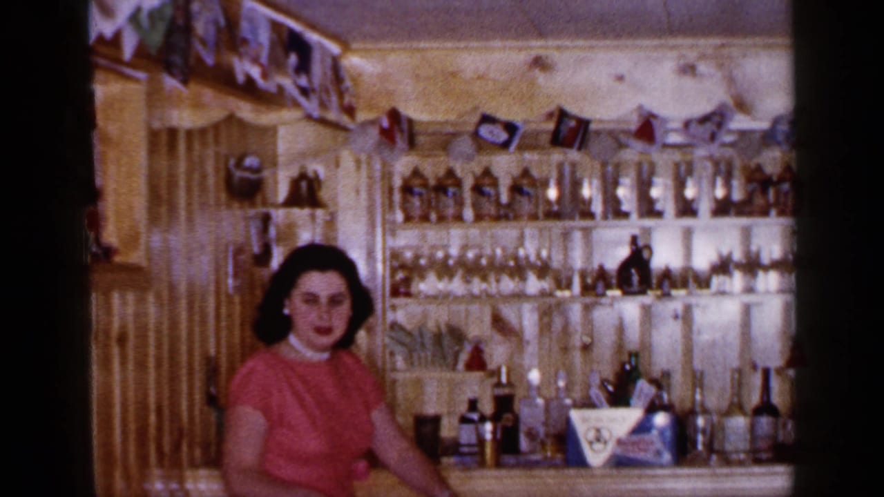 Woman seated beside a bar counter with shelves of glasses, bottles, and hanging pennants in the background.