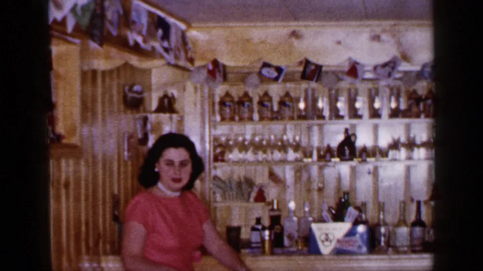 A woman sits near a home bar in Long Island, New York, with glassware, bottles, and party pennants visible in the 1965 interior.