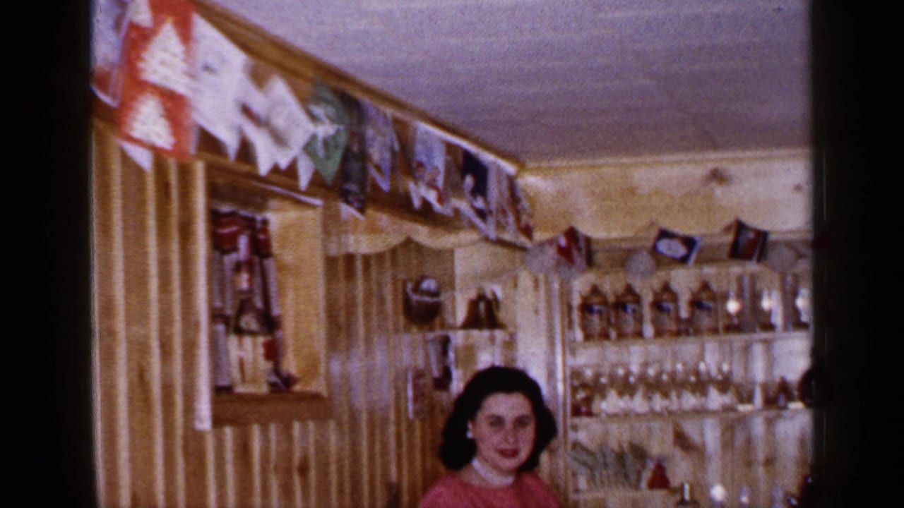 The camera tilts or reframes to show more of the home bar, with a seated woman below shelves of bottles, glassware, and decorations.