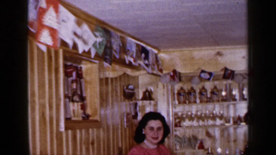 The camera tilts or reframes to show more of the home bar, with a seated woman below shelves of bottles, glassware, and decorations.