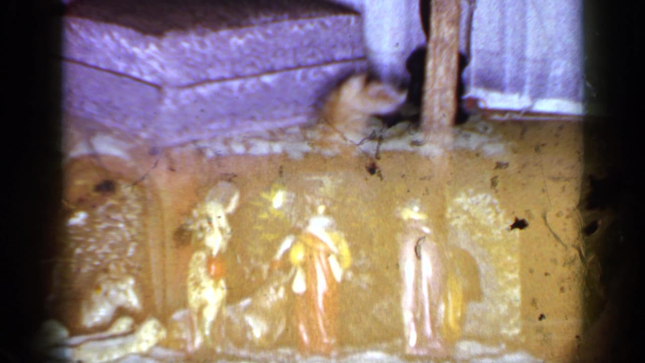 The cat appears near a small nativity display on the floor, with carpet, furniture, and curtains in the 1965 home setting.