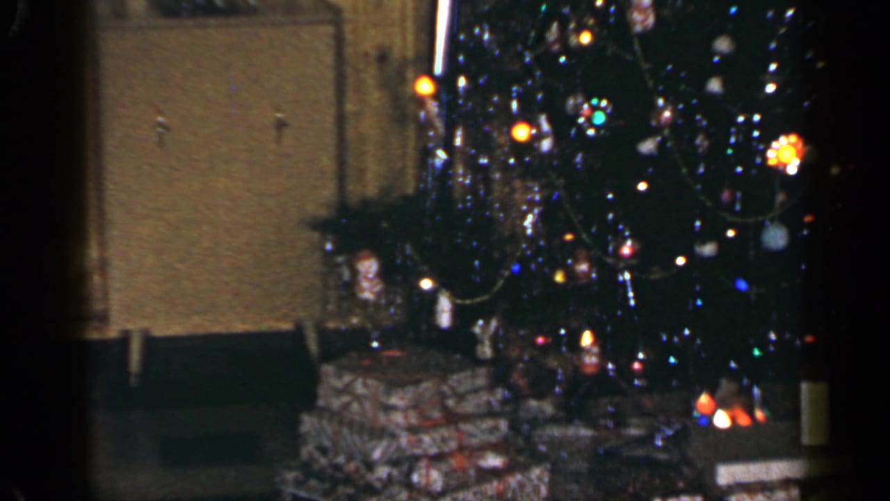 1965 Long Island home-movie frame emphasizing the lit Christmas tree, ornaments, tinsel, and presents along the lower edge.
