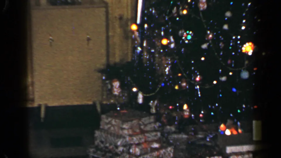 1965 Long Island home-movie frame emphasizing the lit Christmas tree, ornaments, tinsel, and presents along the lower edge.