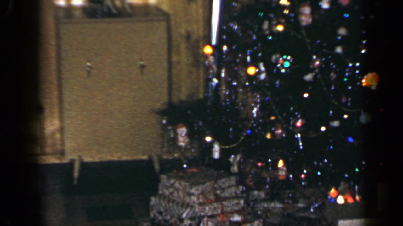 1965 Long Island home-movie frame of a lit Christmas tree beside wrapped presents and a cabinet in a wood-paneled living room.