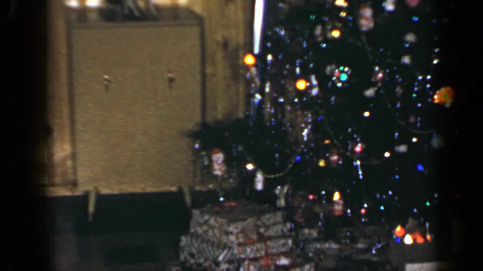 1965 Long Island home-movie frame of a lit Christmas tree beside wrapped presents and a cabinet in a wood-paneled living room.