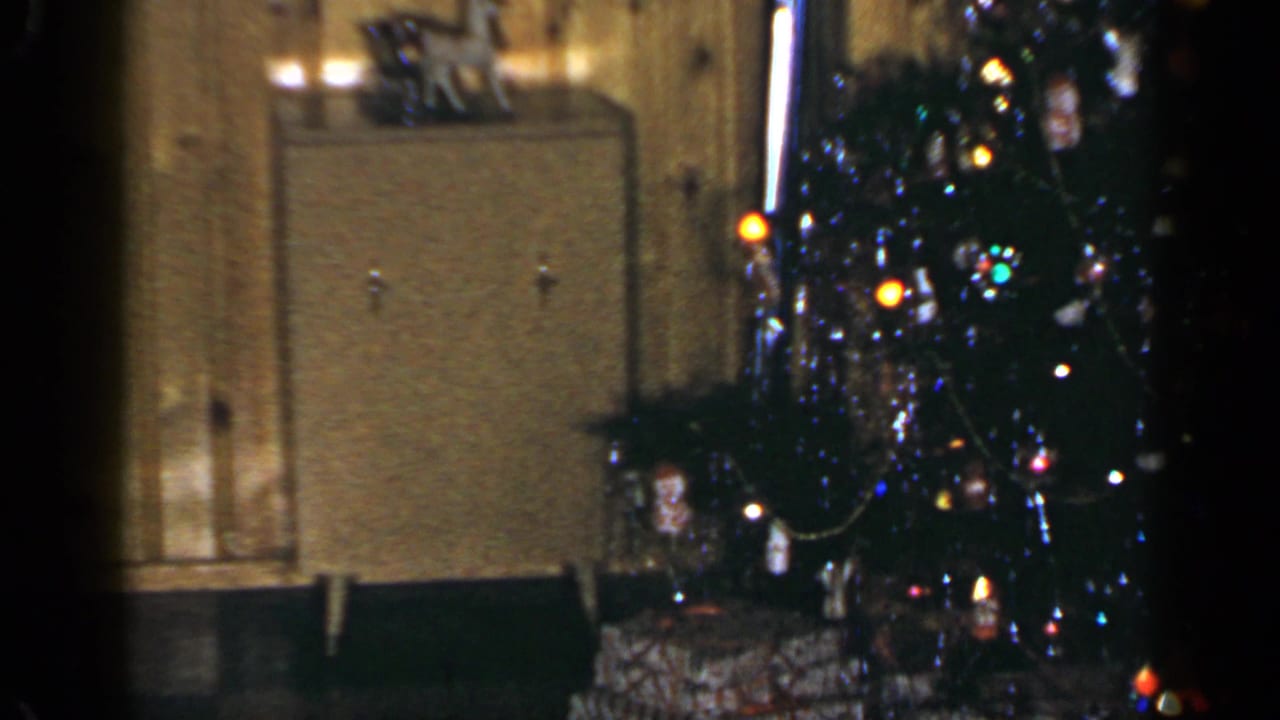 Christmas tree and presents beside a cabinet in a dim living room.