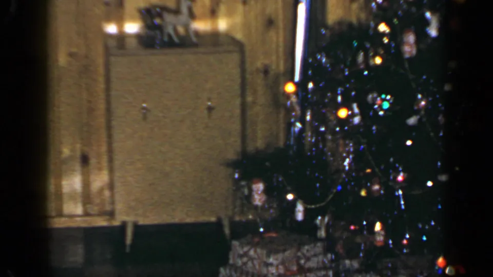 1965 Long Island interior view with a decorated Christmas tree, wrapped gifts, and cabinet against wood-paneled walls.