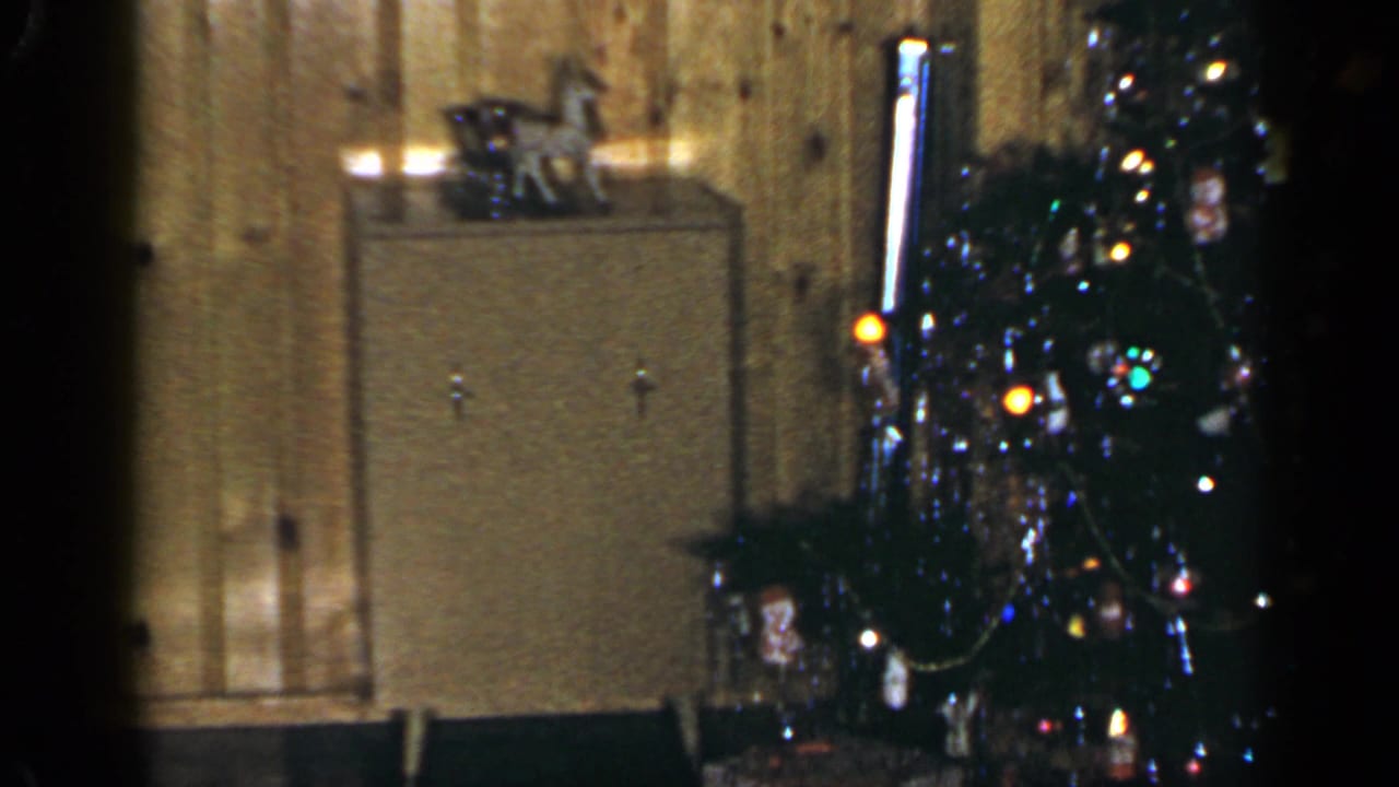 1965 Long Island home interior showing a cabinet, lit Christmas tree, ornaments, and wrapped presents along a paneled wall.