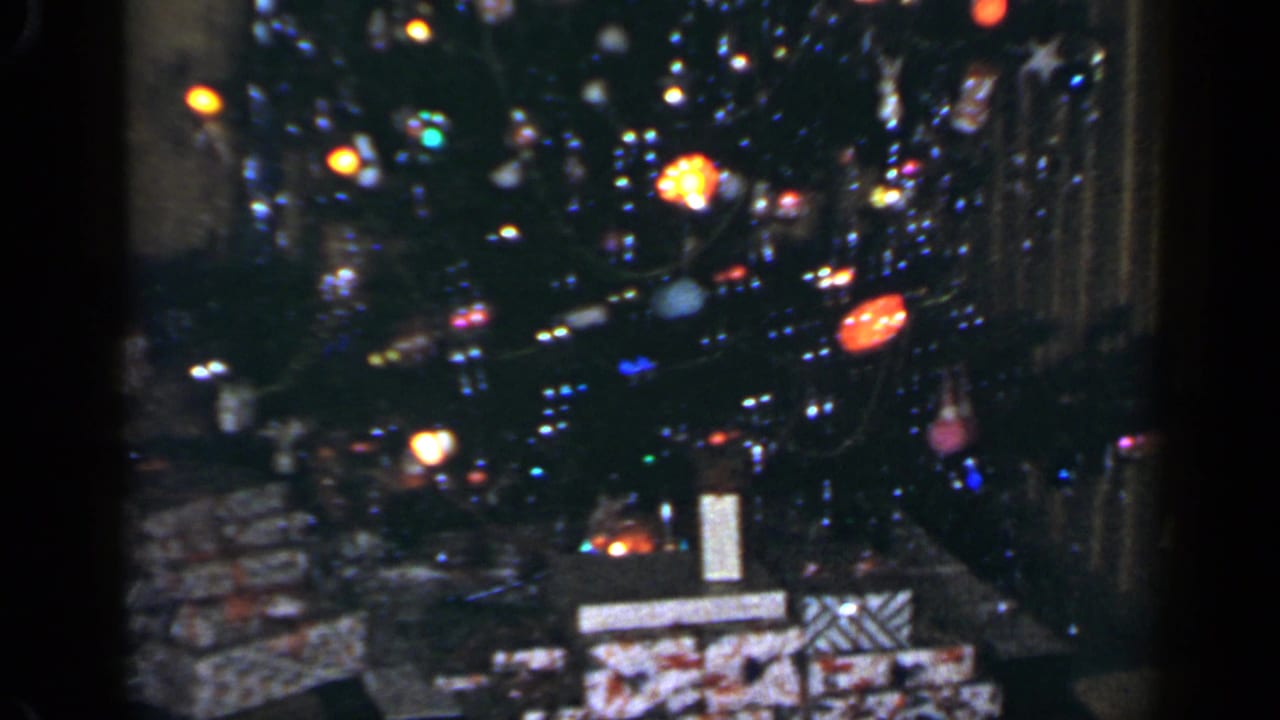 Close 8mm view of a 1965 Long Island Christmas tree with glowing lights, ornaments, tinsel, and gifts underneath.