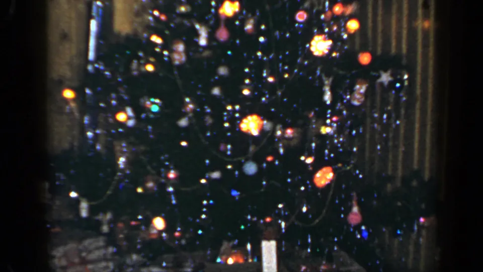1965 Long Island home-movie frame showing a lit decorated Christmas tree in a wood-paneled interior.