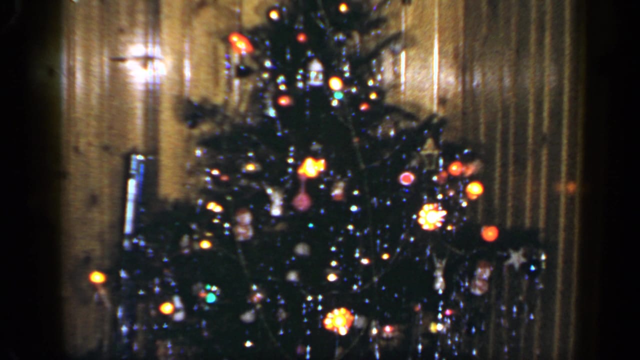 1965 home-movie frame of a lit Christmas tree and wood-paneled room, with ornaments and tinsel visible.