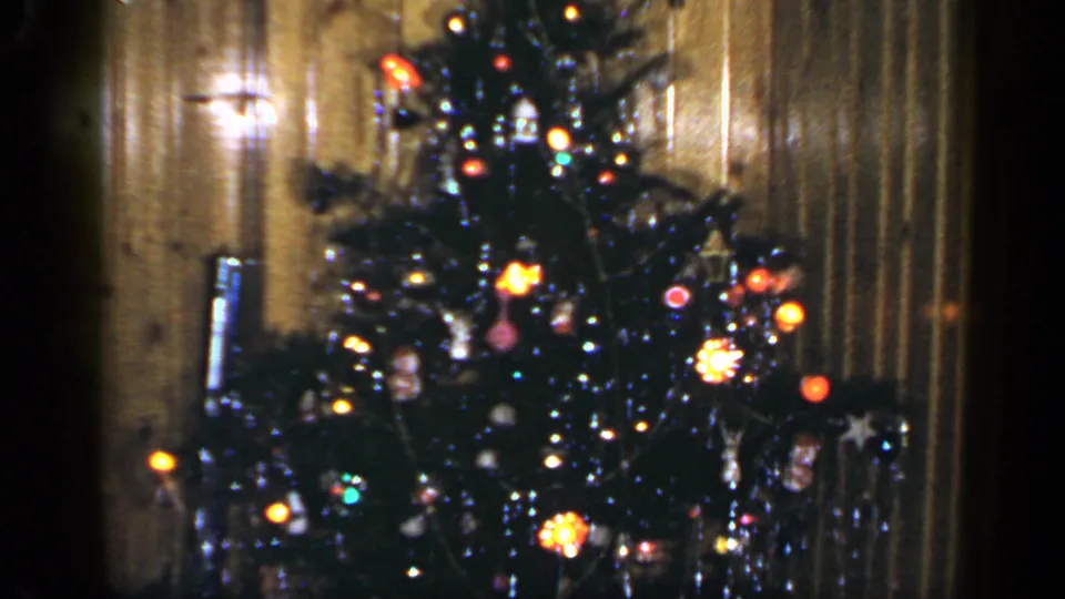 1965 home-movie frame of a lit Christmas tree and wood-paneled room, with ornaments and tinsel visible.