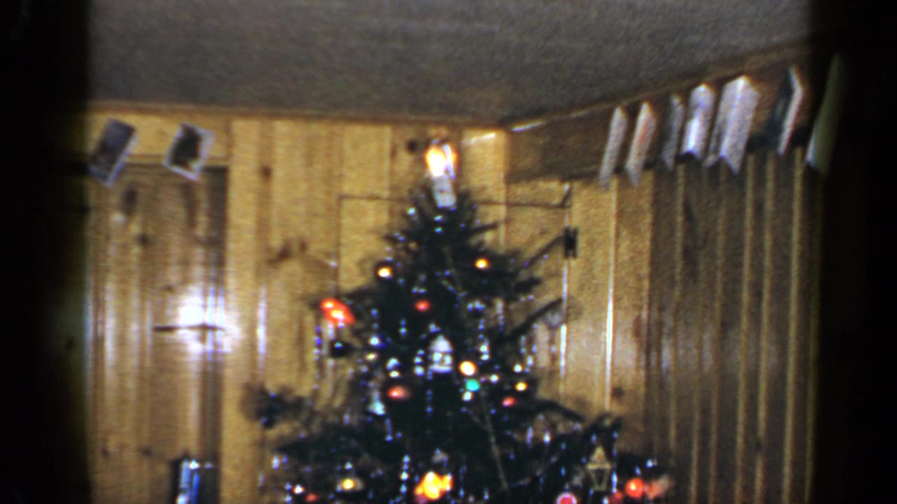 1965 Long Island home-movie frame of a decorated Christmas tree with lights, curtains, and hanging cards overhead.