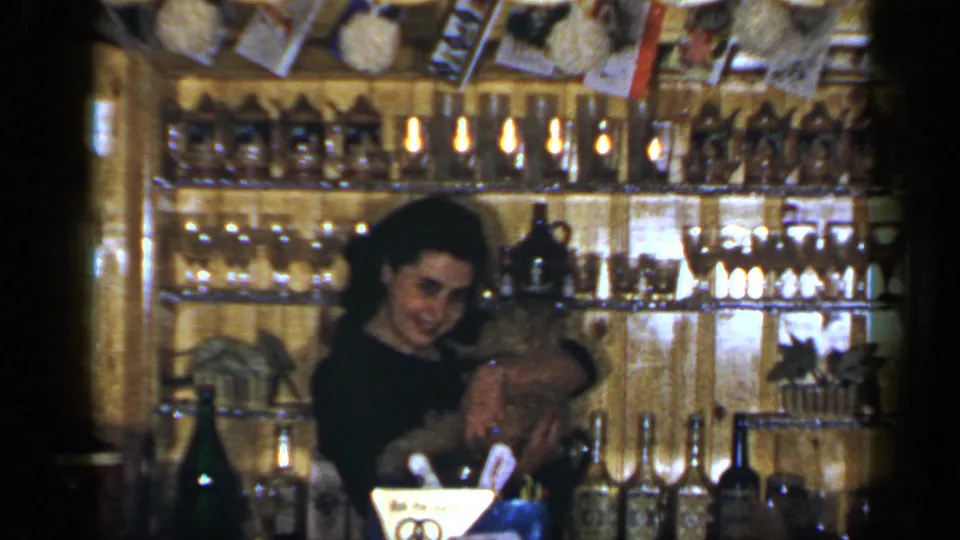 A smiling woman holds a small dog in a cozy Long Island shop interior, with wood shelves, bottles, glassware, and decorations behind her.