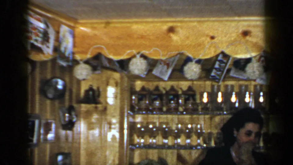 A Long Island 1965 home-movie frame shows the woman partly cropped beside a wood-paneled bar area filled with glasses, bottles, photos, and trim.