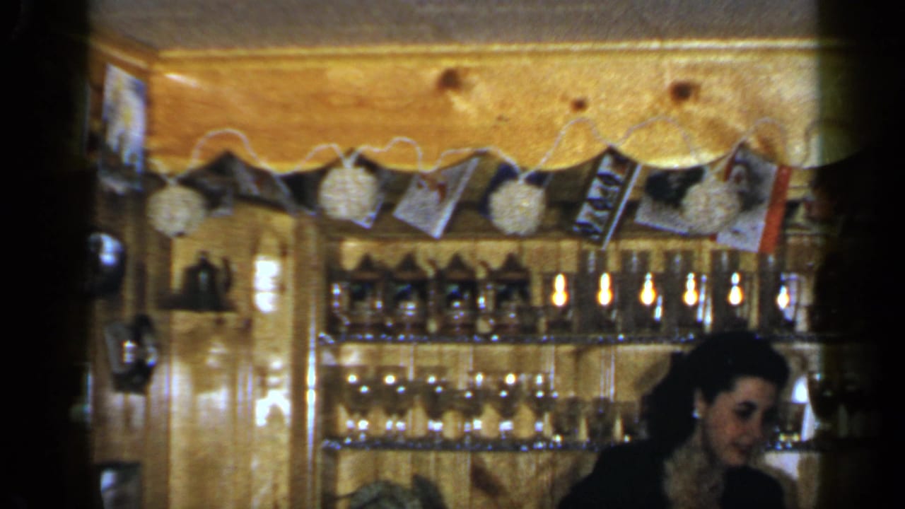 Woman behind a wooden bar with bottles, glass shelves, and a hanging photo garland.