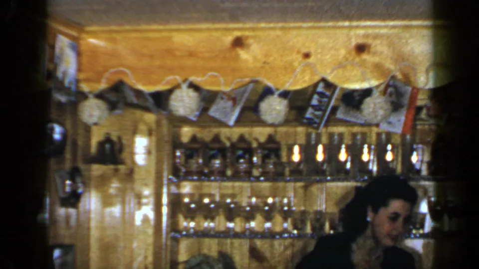 1965 Long Island 8mm frame of a woman behind a warm wooden bar with glassware, bottles, photos, and white pom-poms above.