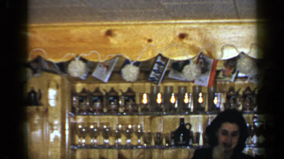 1965 Long Island bar interior with the woman near the right edge, shelves of glassware, bottles, and a decorated photo garland overhead.