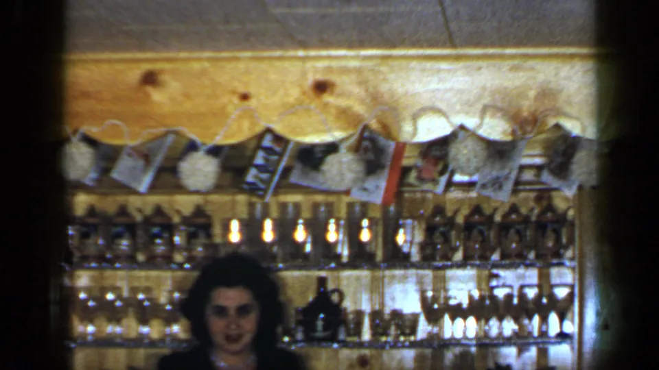 1965 Long Island home-movie frame showing the woman centered below shelves of glasses and bottles, with photos and pom-poms above.