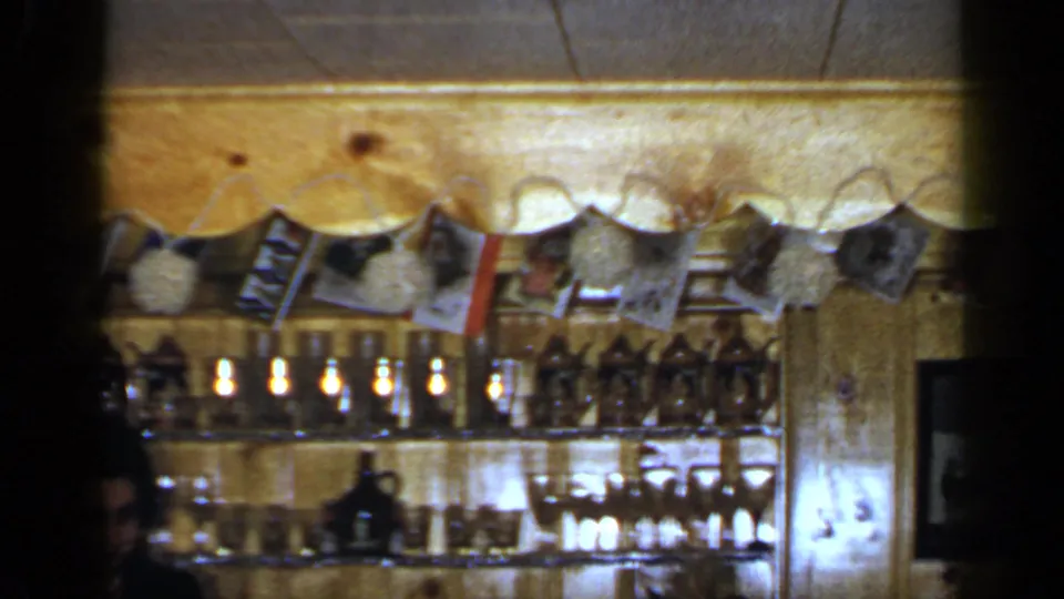 Final 1965 Long Island 8mm frame of the wooden bar area, with glassware, bottles, hanging photographs, and the woman partly obscured.