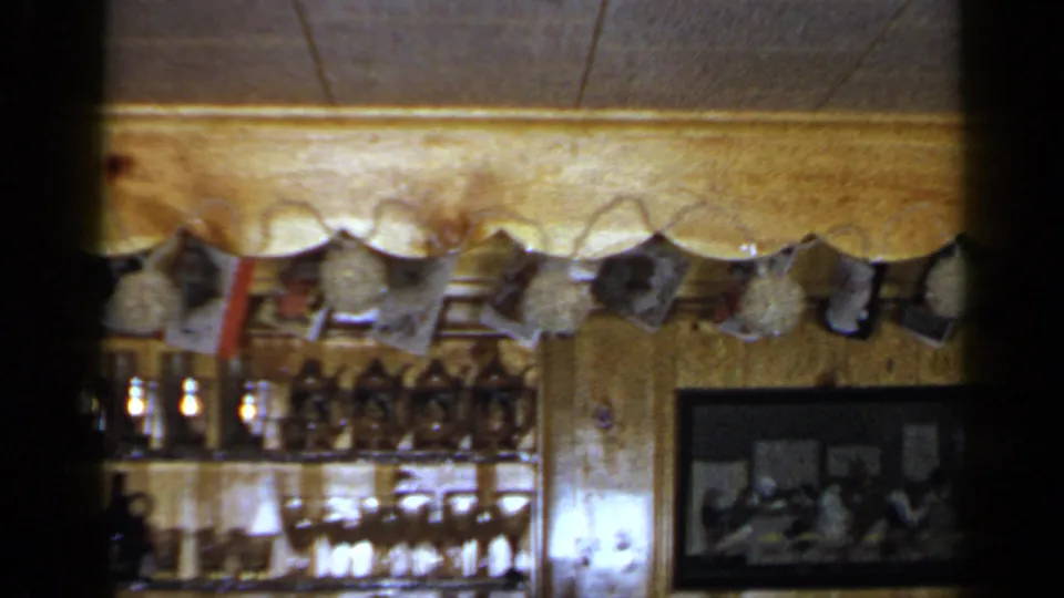 1965 Long Island home interior with scalloped trim, hanging snapshots or postcards, a glassware shelf, and a framed group photograph.
