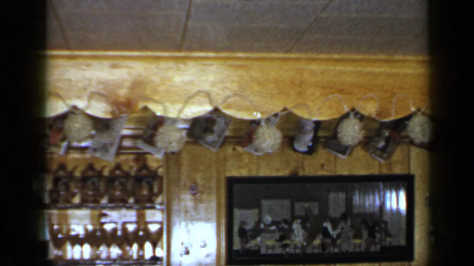 1965 Long Island frame showing hanging small prints, wood paneling, partial glassware shelves, and a framed group photograph in a private room.