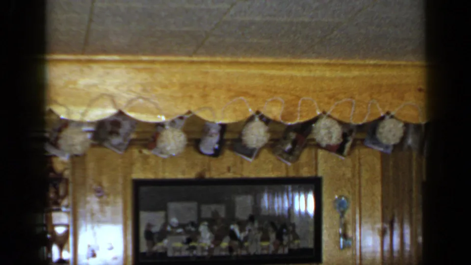 1965 Long Island home-movie frame of a paneled interior with hanging pictures, a framed group photograph, scalloped trim, and a small wall ornament.