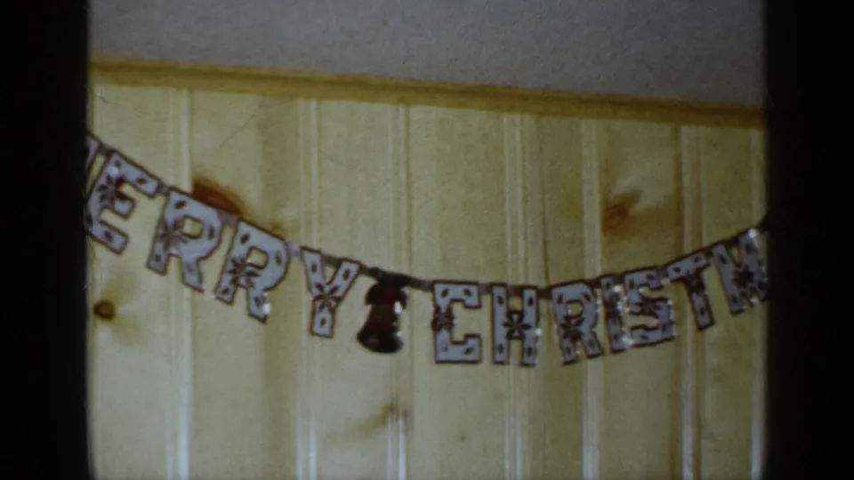 1965 Long Island home-movie frame showing the lower arc of a Merry Christmas banner over a warm wood-paneled wall.
