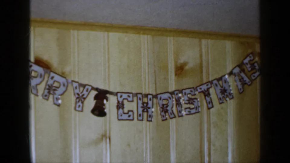 1965 Long Island frame showing a Merry Christmas decoration spanning a pale wood-paneled wall in a private interior.