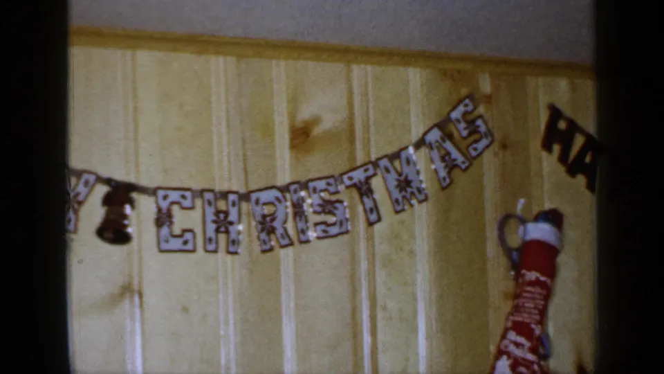1965 Long Island home-movie frame with the Merry Christmas banner centered and a red stocking hanging at the right edge.