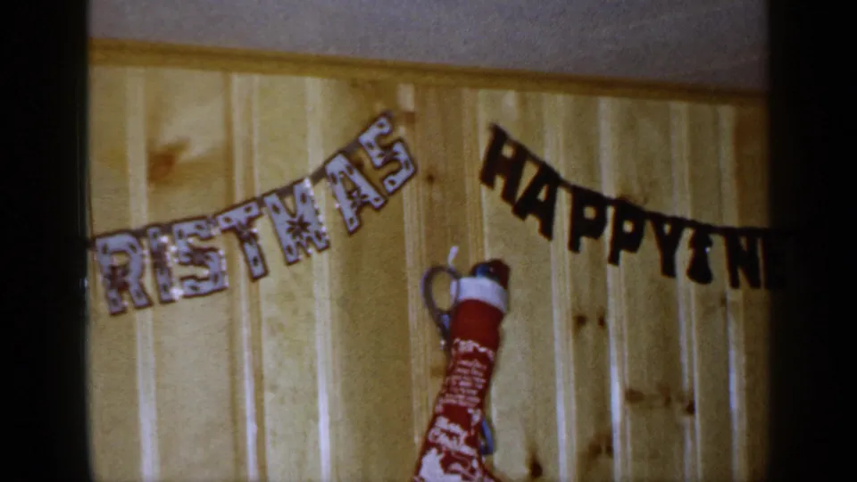 1965 Long Island home-movie frame with a red stocking below overlapping Christmas and Happy New Year holiday banners.